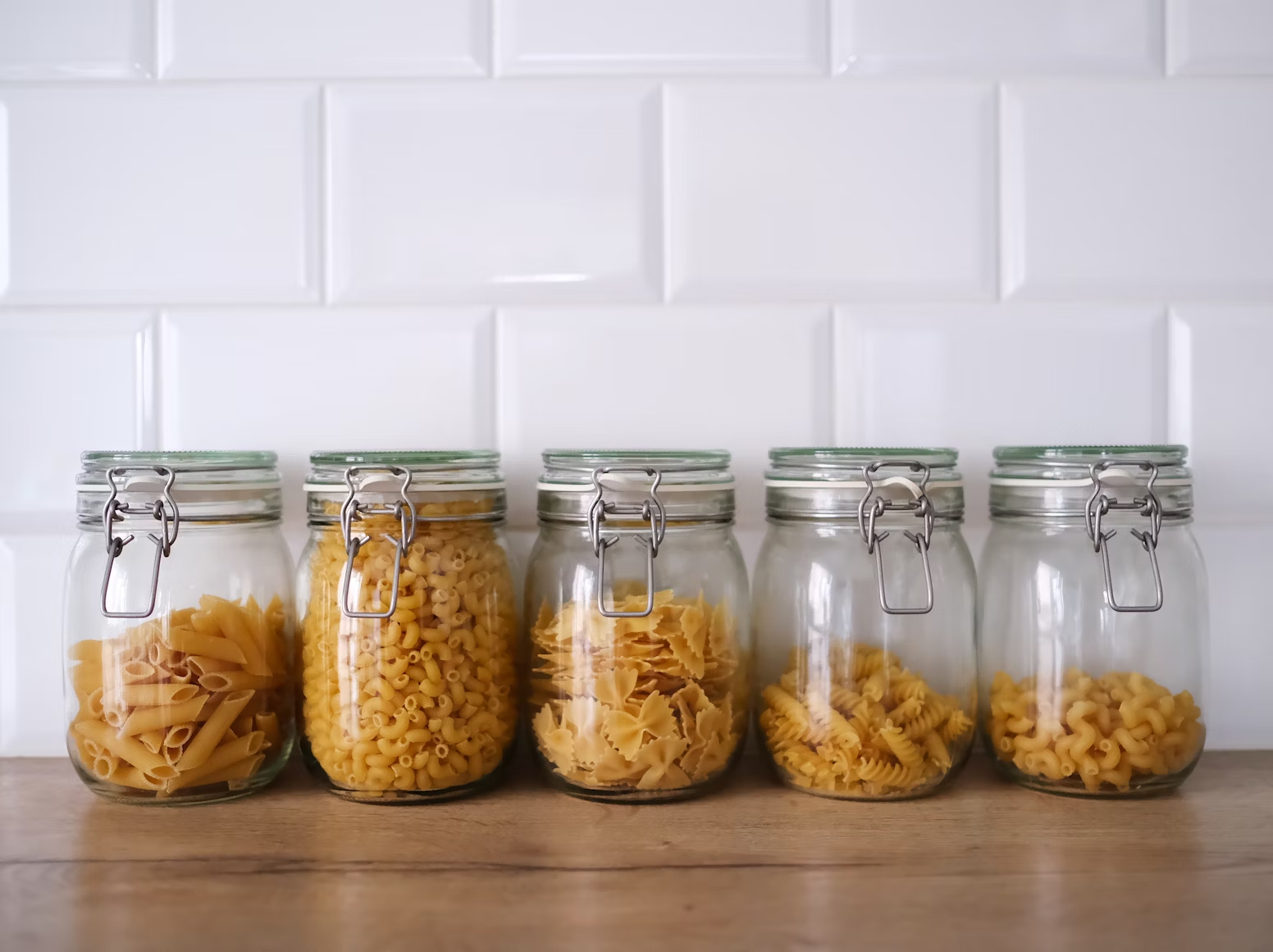 Glass jars filled with various pasta shapes organized on a wooden shelf